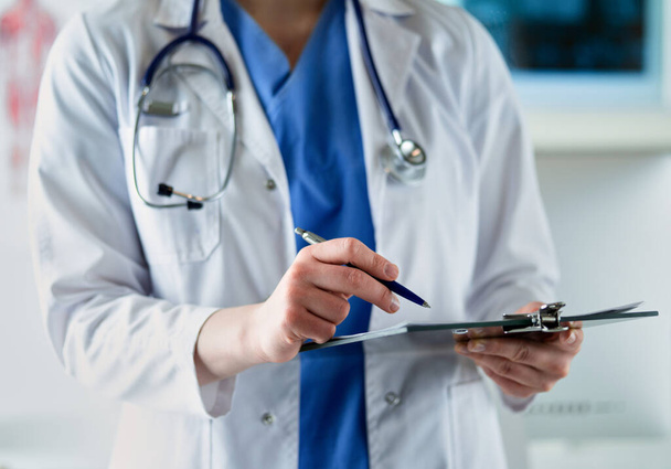 Doctor with a stethoscope, holding a notebook in his hand. Close-up of a female doctor filling up medical form at clipboard while standing straight in hospitalDoctor with a stethoscope, holding a - Fotoğraf, Görsel