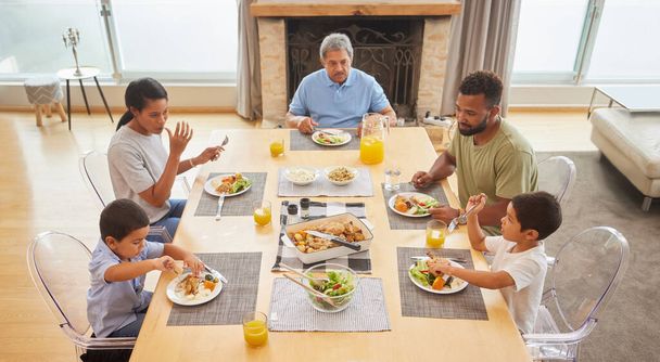 Vista aérea de una familia de raza mixta sentada en una mesa almorzando en el salón de su casa. Abuelos hispanos comiendo con sus hijos y nietos en casa. - Foto, Imagen