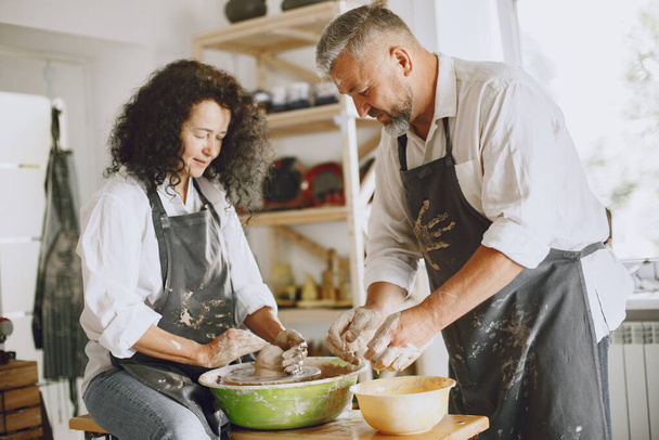 Mutual creative work. Adult elegant couple in casual clothes and aprons. People creating a bowl on a pottery wheel in a clay studio. - Foto, imagen