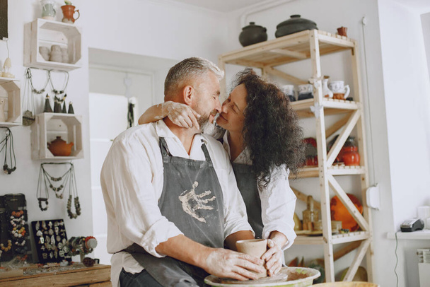 Mutual creative work. Adult elegant couple in casual clothes and aprons. People creating a bowl on a pottery wheel in a clay studio. - Photo, image