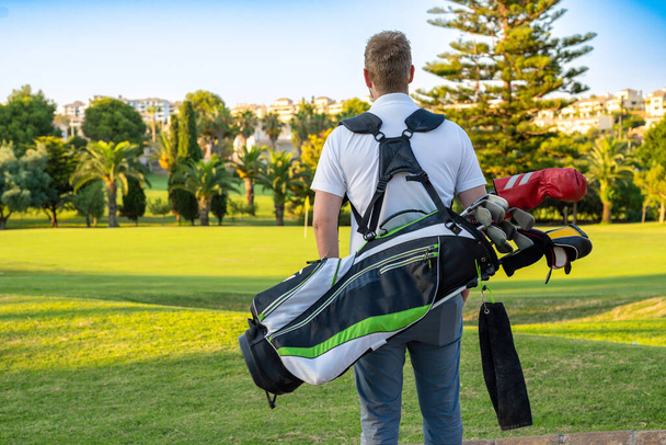 Widok z tyłu młodego przystojnego mężczyzny na polu z torbą golfową. - Zdjęcie, obraz
