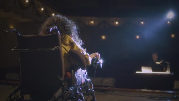 Eine behinderte Schauspielerin mit spinaler Muskelatrophie im Rollstuhl auf einer von einem Scheinwerfer beleuchteten Bühne im Gespräch mit dem Regisseur im Theater - Filmmaterial, Video