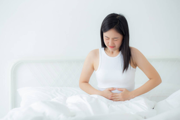 Jonge Aziatische vrouw zittend op bed pijn maag of darm met ziekte constipatie in de slaapkamer thuis, ongelukkige vrouwelijke ziek en menstruatie en buikpijn, medische en gezondheidsconcept. - Foto, afbeelding