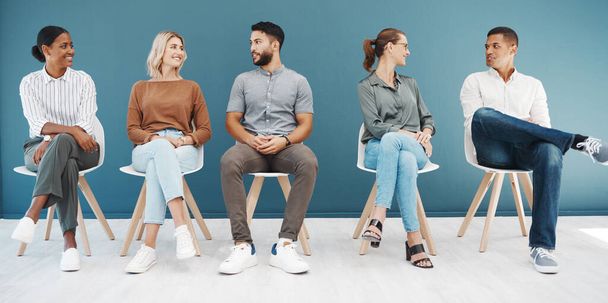 Diverse group of confident business men and women sitting on chairs in waiting room against a blue background. Happy hopeful candidates and applicants ready for shortlist interview in line for job - Zdjęcie, obraz