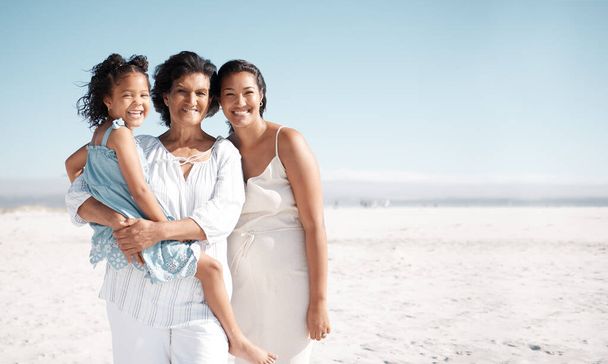 Smiling mixed race family standing together on a beach with copyspace. Happy hispanic grandmother bonding with granddaughter over weekend. Adorable little girl enjoying free time with single mother. - Photo, Image