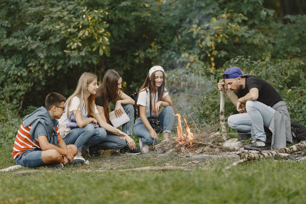 Adventure, travel, tourism, hike and people concept. Group of smiling friends in a forest. People sitting near bonfire. - Photo, Image