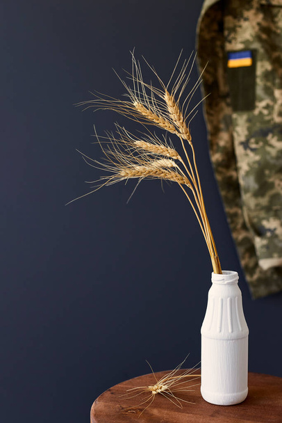 Bundel tarwe spikes met Oekraïense vlag lint op militaire uniform als achtergrond. Concept van voedselvoorzieningscrisis en wereldwijde voedselschaarste als gevolg van oorlog in Oekraïne. - Foto, afbeelding
