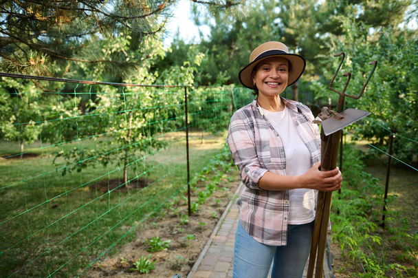 Charming Caucasian woman, amateur farmer, successful female gardener smiles to camera with gardening tools in her hands, standing in a countryside organic farm. Horticulturist portrait, agribusiness - Foto, afbeelding