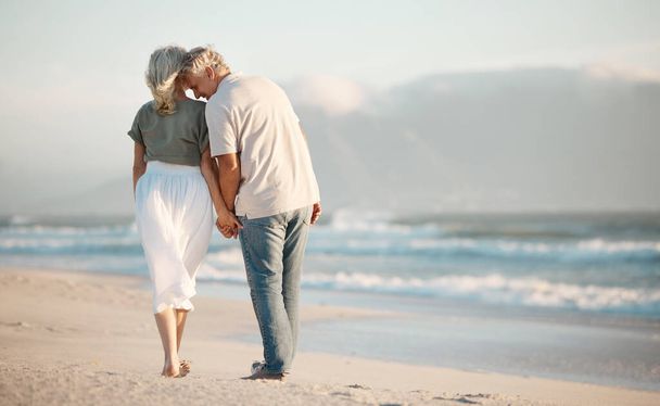 Back of a senior couple walking on a beach together. Senior couple holding hands and walking on the beach. Mature couple enjoying a vacation together. Happy husband being affectionate with his wife. - Foto, Imagen