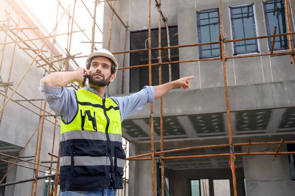 Young attractive construction smiling bearded using smartphone calling to client, worker in vest with white helmet standing on construction site - Fotografie, Obrázek