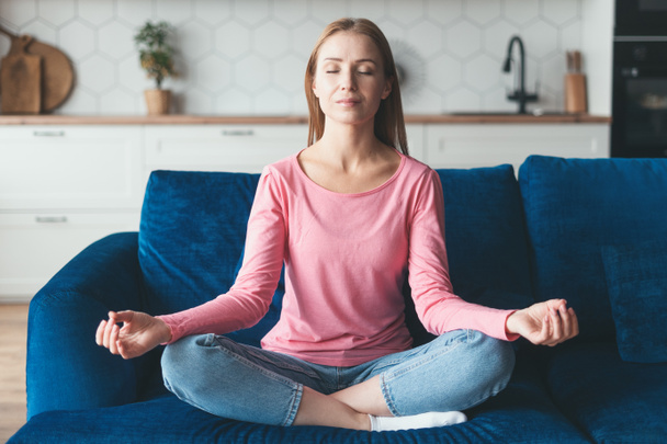 Mental health, mindfulness lifestyle, anxiety relief. Calm woman sitting on couch in lotus pose, doing yoga exercise at home, put hands on lap folded fingers in mudra gesture and enjoy meditation - Фото, зображення