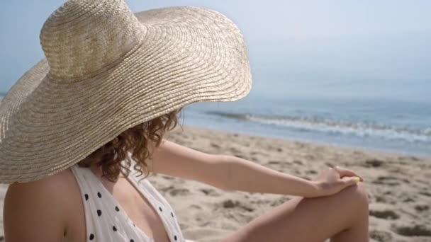 Потрясающая молодая женщина, смотрящая в камеру, соблазнительно расслабляется на солнечном побережье вблизи. Портрет великолепной кудрявой модели, позирующей на песчаном пляже в широкой шляпе с полями. Сексуальная девушка сидит рядом с океаном. - Кадры, видео
