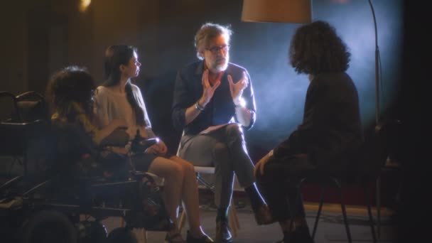 Eine Gruppe von Schauspielern mit einer behinderten Schauspielerin und einem Regisseur mit Drehbuch sitzt auf einer dunklen Bühne und diskutiert gemeinsam eine Theateraufführung - Filmmaterial, Video