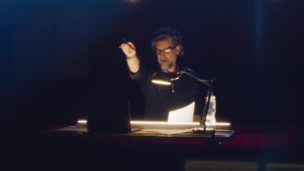 Männlicher Regisseur mit Drehbuch gibt Schauspielern Anweisungen, während er während der Probe in einem dunklen Theater mit Lampe am Tisch sitzt - Filmmaterial, Video