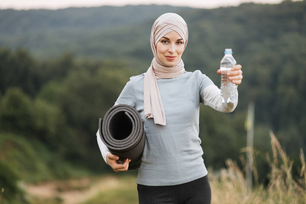 Front view of charming woman in hijab and sport clothes standing at green park and holding yoga mat and bottle of water. Concept of people, activity and recreation. - Foto, afbeelding