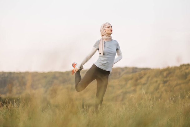 Active woman in sport clothes and hijab stretching legs at summer park. Young arabian girl standing on background of beautiful park at morning and doing physical exercises Sport activity outdoors. - Fotografie, Obrázek