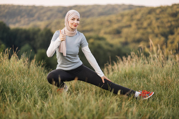 Muslim, arab, islamic pretty slim woman in sporty outfit and hijab stretching her leg during morning workout showing thumb up in park outdoors. Healthy lifestyle concept. - Foto, afbeelding