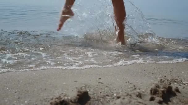 Unknown barefoot girl splashing water stepping ocean waves close up. Beautiful slim tanned legs walking wet sea sand summer day. Happy joyful woman have fun relaxing on seacoast. Vacation concept. - Video, Çekim