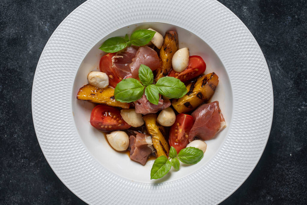 Grilled peach salad with mozzarella, prosciutto ham, red tomato, green basil and soy sauce. Fresh gourmet salad in white plate on black background, close up, top view - Photo, Image