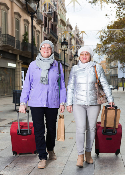 Happy traveling elderly women strolling with luggage along city street - Fotó, kép