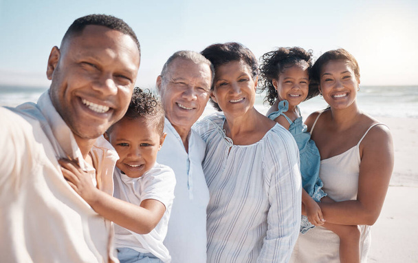 Portrait of smiling mixed race family with little boy and girl taking selfie while standing together on beach. Adorable little son and daughter bonding with mother, father, grandmother and grandfather - 写真・画像