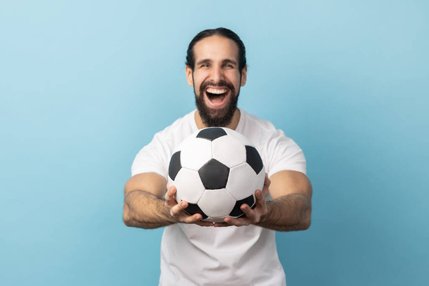 Portrait of man with beard wearing white T-shirt holding out soccer ball in hand and looking at camera with excited facial expression. Indoor studio shot isolated on blue background. - Photo, Image