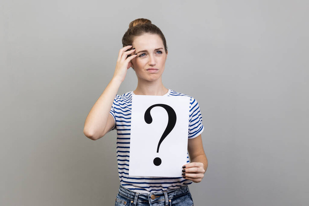 Portrait of pensive thoughtful woman wearing striped T-shirt looking away, holding paper with question mark, thinks about solving problem. Indoor studio shot isolated on gray background. - Foto, imagen