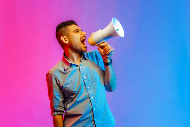Portrait of aggressive man in shirt screaming at megaphone, making announce, protesting, looking away with angry expression. Indoor studio shot isolated on colorful neon light background. - Foto, Bild