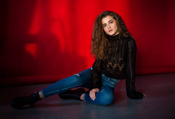 Attractive young woman with makeup and curly hair, dressed black blouse and jeans, isolated red background. - Φωτογραφία, εικόνα