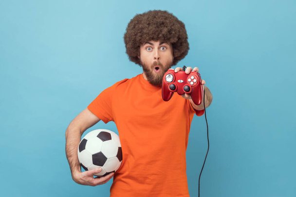 Portrait of excited amazed man with Afro hairstyle wearing orange T-shirt holding soccer ball and showing red joypad to camera, video game. Indoor studio shot isolated on blue background. - Photo, Image