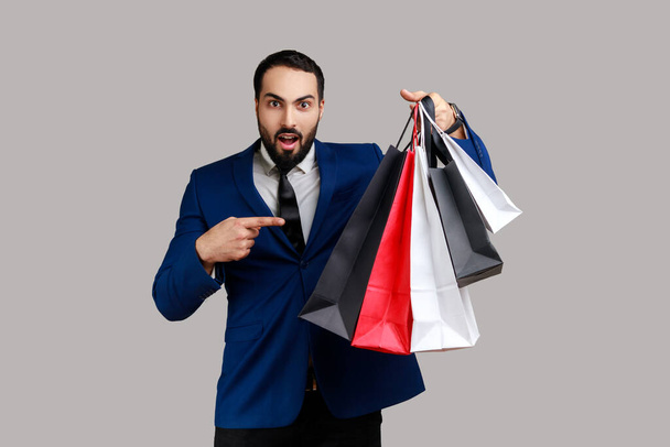 Bearded businessman pointing finger at paper bags in her hand, shocked with low prices for good quality, wearing official style suit. Indoor studio shot isolated on gray background. - 写真・画像