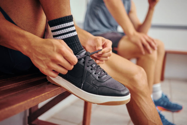Squash player tying his shoe lace. Hands of a fit athlete getting ready for a squash match. Closeup of a player tying the laces of his sport sneaker. Player sitting on a bench with his friend - Photo, Image