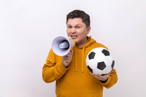 Portrait of middle aged man holding soccer black and white ball and megaphone, making announce on football match, wearing urban style hoodie. Indoor studio shot isolated on white background. - Foto, afbeelding
