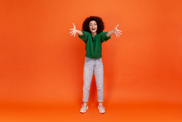 Come to my arms! Full length of friendly young adult woman with Afro hairstyle wearing green casual style sweater spreading hands, inviting, embracing. Indoor studio shot isolated on orange background - 写真・画像