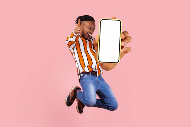 Hey you look here. Happy young man flying and jumping in air and showing big mobile empty screen for copy space and advertising area. indoor studio shot isolated on pink background - Valokuva, kuva