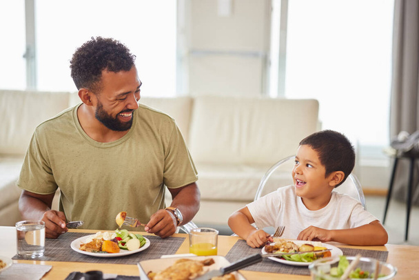 Closeup of a mixed race male and his son enjoying some food at the a table during lunch at home in the lounge. Hispanic father smiling and eating alongside his son at home. - Photo, image