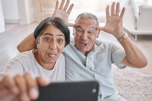 Closeup of an affectionate senior couple taking selfies while relaxing in the living room at home. Mixed race couple having fun taking photos in the lounge at home. - Φωτογραφία, εικόνα