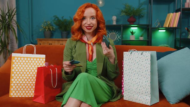 幸せなお店の消費者の赤毛の若い女性が自宅でオンライン注文を行うショッピングバッグに座って、クレジットプラスチック製の銀行カードでお支払い。インターネットの概念の割引販売。ジンジャー女の子オンソファ - 写真・画像