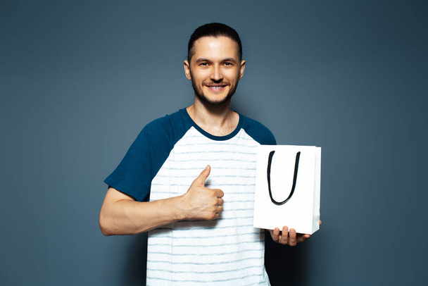 Studio portrait of young smiling man, holding a white bag and showing thumb up. - Photo, Image