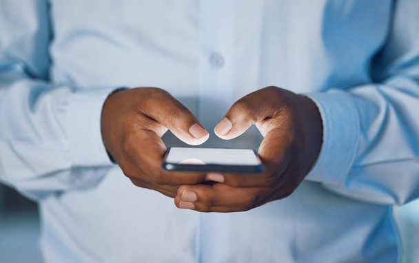 Closeup hands of african man using a phone while standing in his office. African american business man sending a text message while working late at night at his work. Putting in overtime after hours. - Фото, зображення