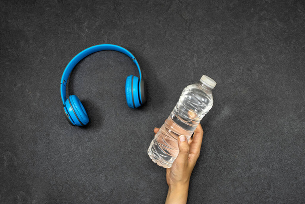 conceito de esporte, Close-up de mão feminina segurando garrafa de água de plástico Backlit com fone de ouvido sem fio azul no fundo grunge - Foto, Imagem