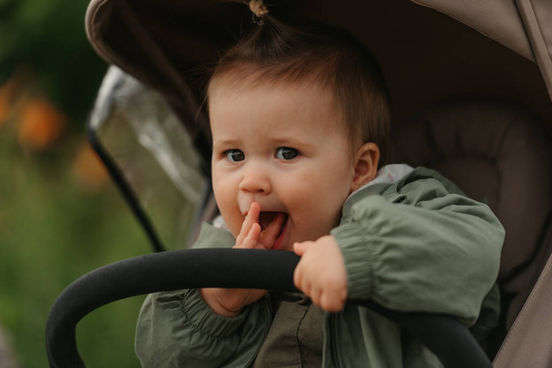 Ein nahes Porträt einer netten Kleinkindfrau, die an einem bewölkten Tag im Kinderwagen die Finger in den Mund legt. In einem grünen Dorf sitzt ein junges Mädchen im Regenmantel im Kinderwagen. - Foto, Bild