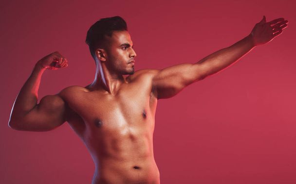 Masculine man standing shirtless with his fist closed and showing fighting technique while posing against a red studio background. Fit indian fighter ready to throw a punch. - Photo, image