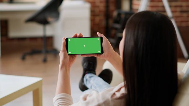 Woman sitting on sofa while holding smartphone with green screen mockup template background. Young adult person with modern touchscreen mobile phone having chroma key isolated display. - Φωτογραφία, εικόνα