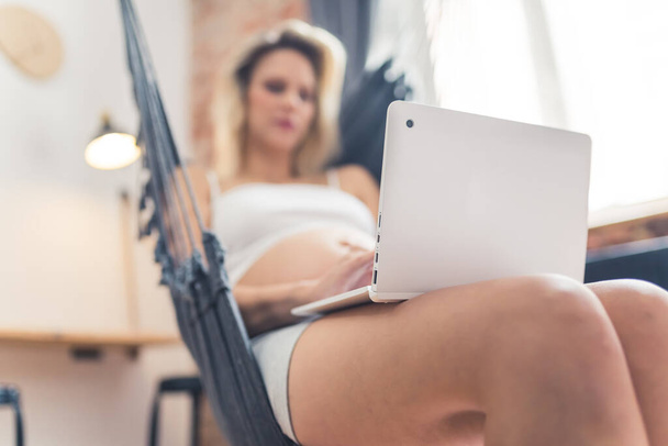 Unscharfe Innenaufnahme einer jungen kaukasischen Schwangeren mit nackten Beinen, die einen Laptop in der Hand hält. Forschung über Arbeit. Hochwertiges Foto - Foto, Bild