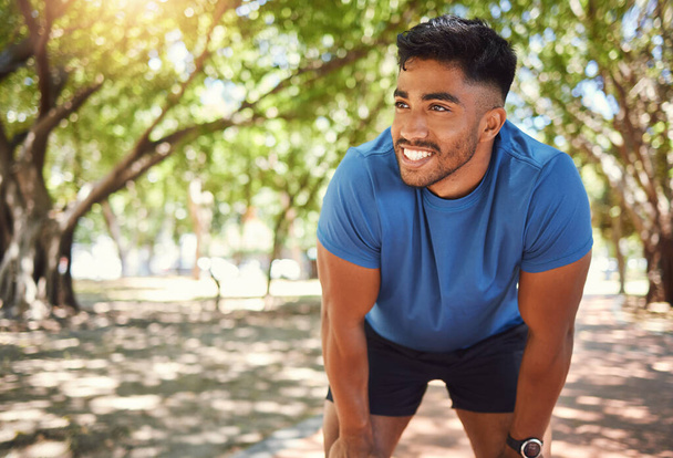 Счастливый атлетичный смешанный расовый человек улыбается стоя с руками на коленях в парке. Латиноамериканец отдыхает после пробежки. Подходящий человек доволен своим выступлением во время упражнений на открытом воздухе. - Фото, изображение