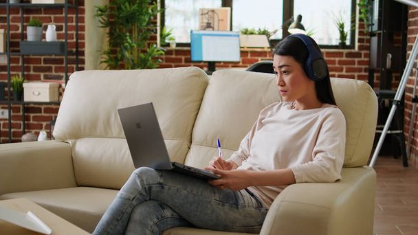 Focused working woman wearing wireless headphones taking notes while doing remote work at home sitting on sofa. Serious looking student attending online class on modern laptop. - Photo, Image