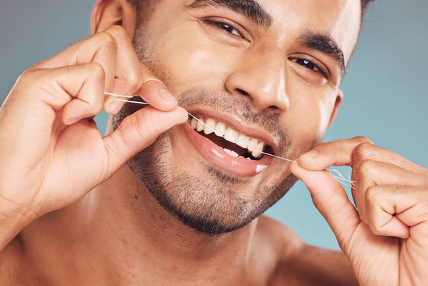 Portrait of one smiling young indian man flossing his teeth against a blue studio background. Handsome guy grooming and cleaning his mouth for better oral and dental hygiene. Floss daily to prevent - Photo, Image