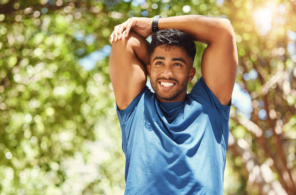 Ajuste atlético hombre de raza mixta sonriendo mientras se estira durante un entrenamiento en el parque. Joven hombre hispano haciendo ejercicios de calentamiento al aire libre en un día soleado. Dedicado al fitness y a un estilo de vida saludable. - Foto, imagen
