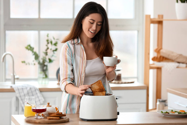 Beautiful young Asian woman with coffee and tasty toasts in kitchen - Foto, afbeelding